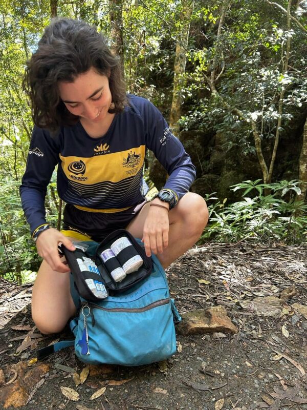 A person crouched in a forest inspecting the contents of an open Hiking First Aid Kit Bundle while on a hiking trip.